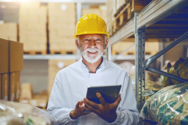 Smiling man in yellow helmet holding tablet in a warehouse.