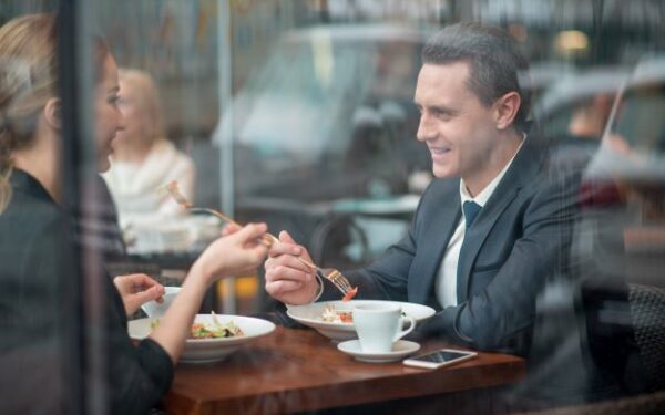 Woman in black blazer sitting across from man in a dark gray blazer eating a meal while in conversation.