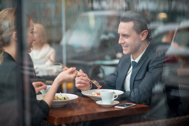 Woman in black blazer sitting across from man in a dark gray blazer eating a meal while in conversation.