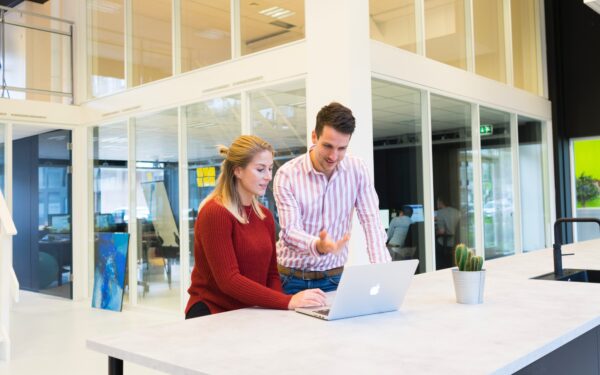 Woman sitting at white desk with man standing on her left gesturing towards a MacBook screen.