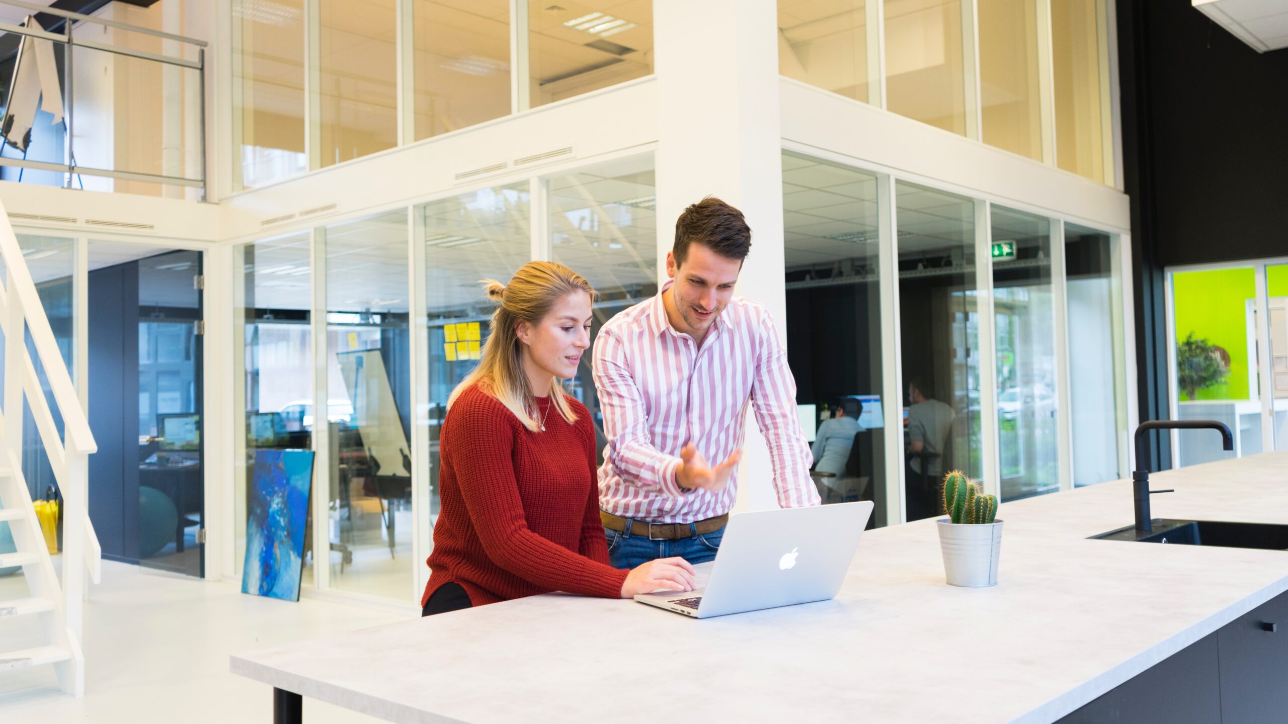 Woman sitting at white desk with man standing on her left gesturing towards a MacBook screen.