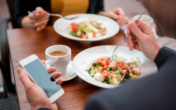 Man in suit jacket holds his cell phone in his left hand while holding a fork sitting across from a colleague.