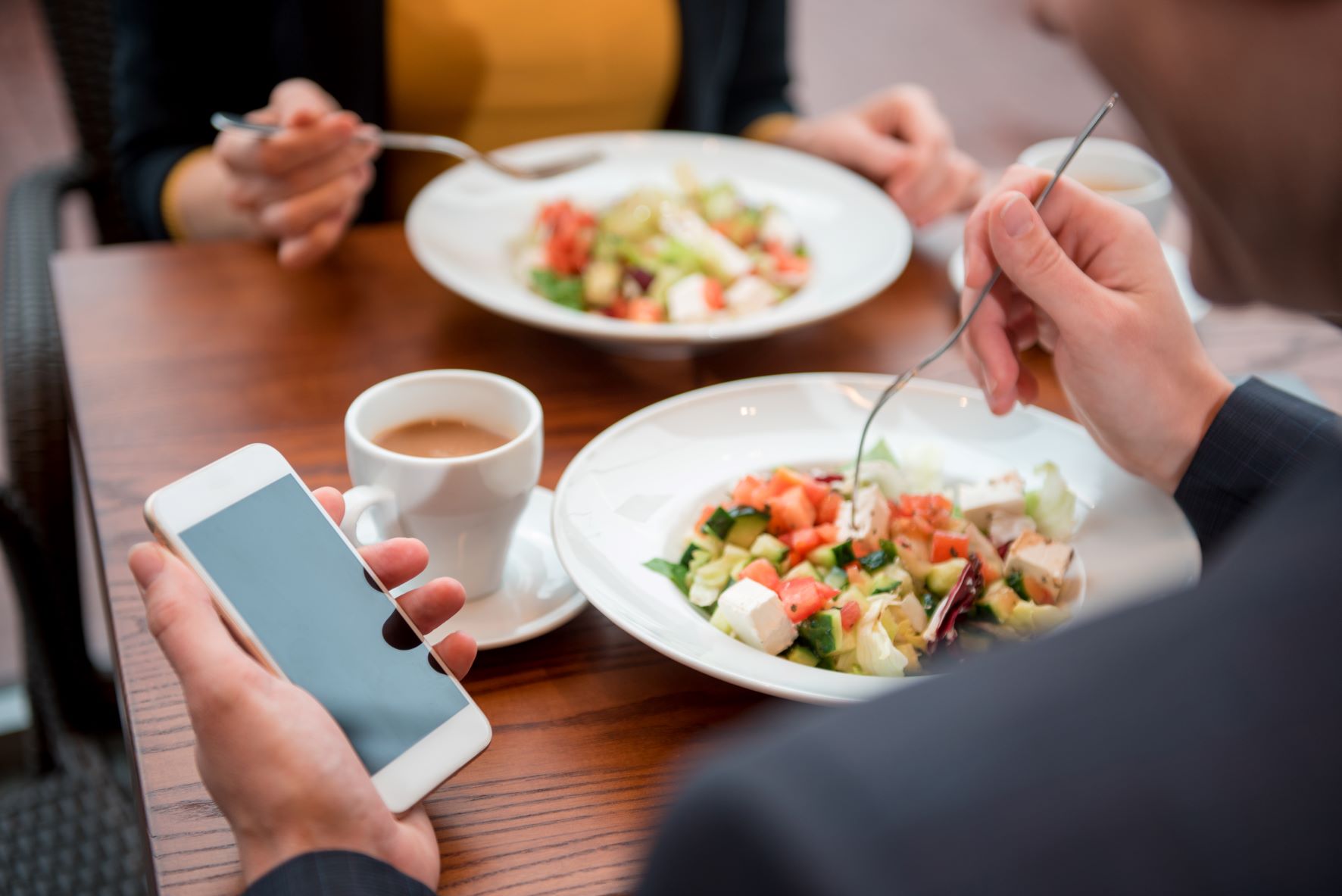 Man in suit jacket holds his cell phone in his left hand while holding a fork sitting across from a colleague.