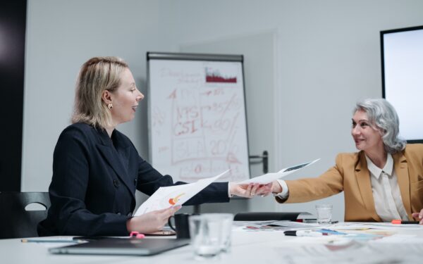 Younger blonde woman handing older woman a paper while sitting at white conference table.