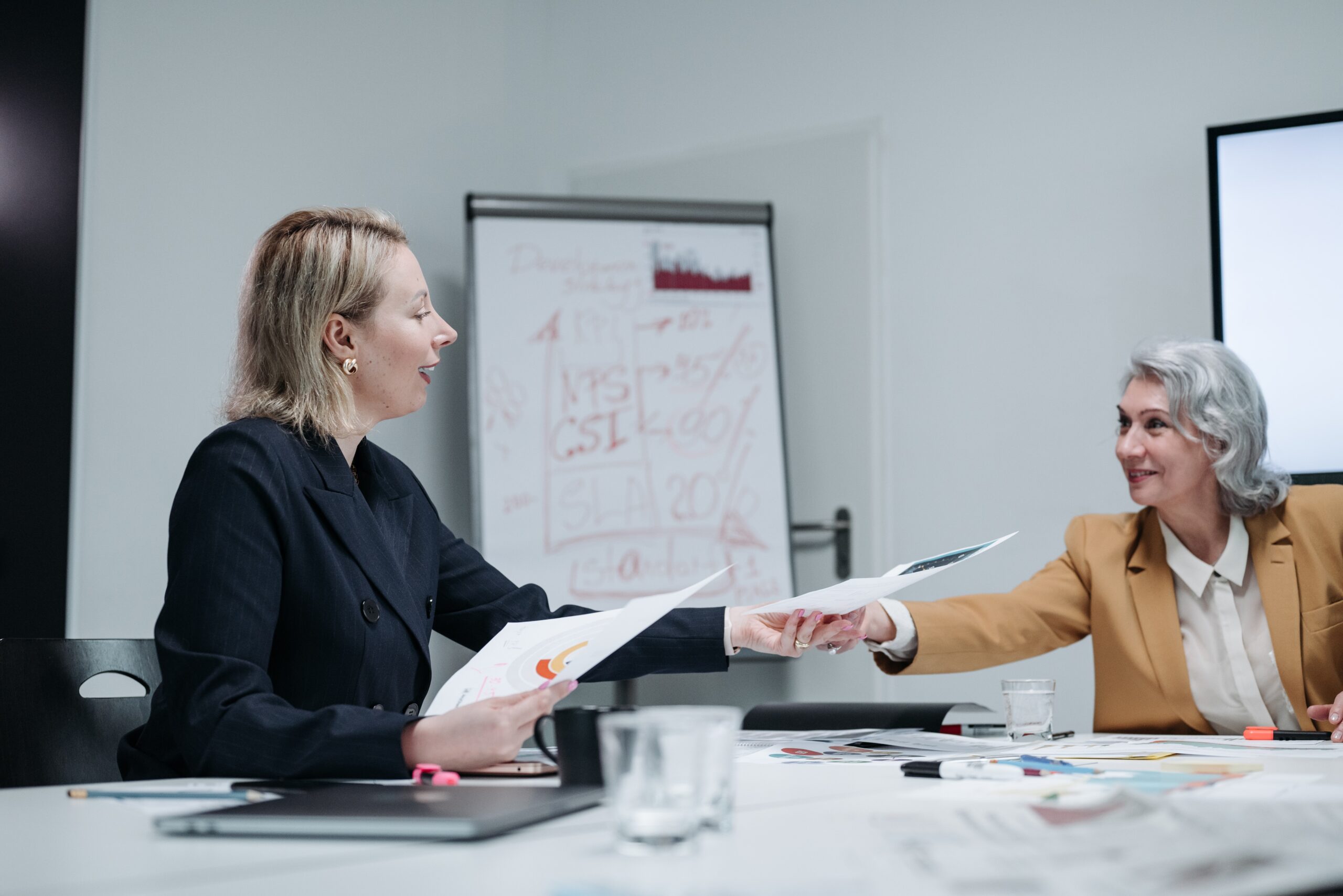 Younger blonde woman handing older woman a paper while sitting at white conference table.