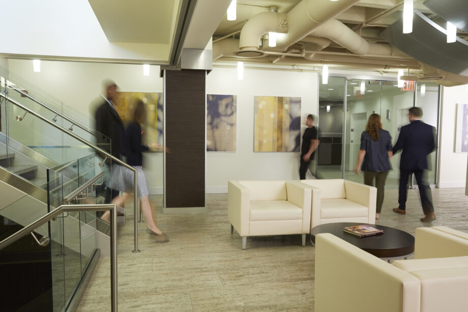 Two people walking down stairs into office lobby while two people walk towards elevators.