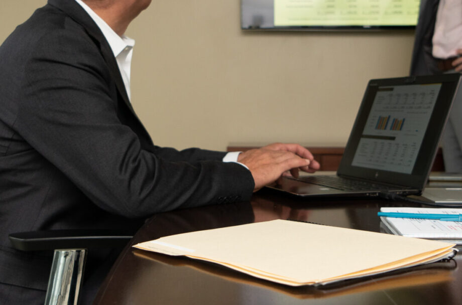 Man in black suit typing on laptop sitting at a wooden conference table.