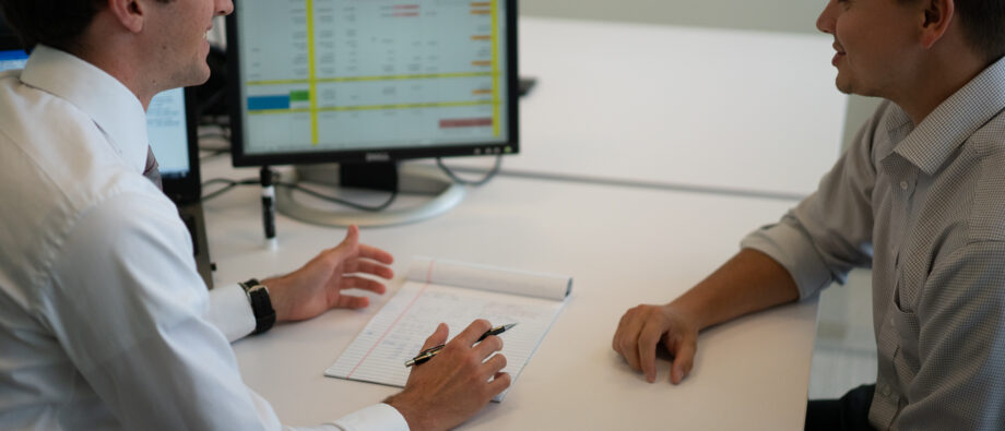 Two men in button up shirts sit at desk discussing a spreadsheet on a computer screen.