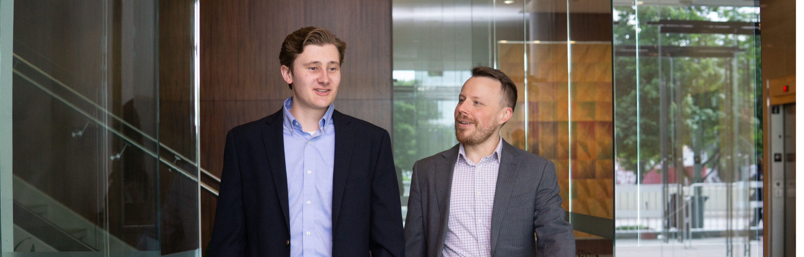 Two men in suits walk in an office building lobby while talking.