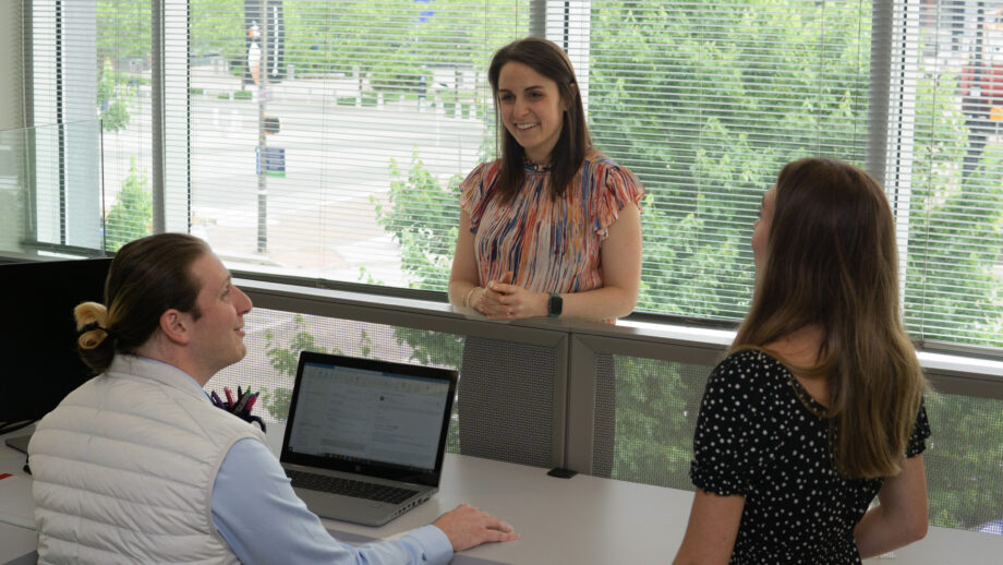 Two women stand at cubicle talking to sitting male coworker.