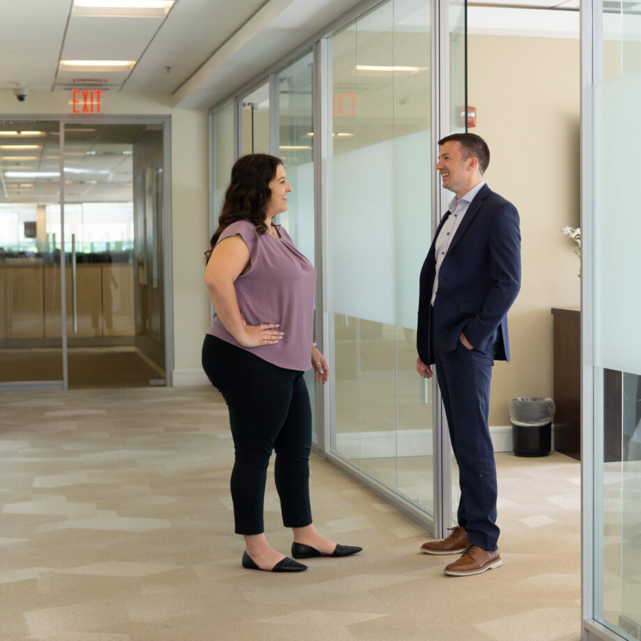 Woman in a purple shirt stands in hallway talking to a man in a blue suit.