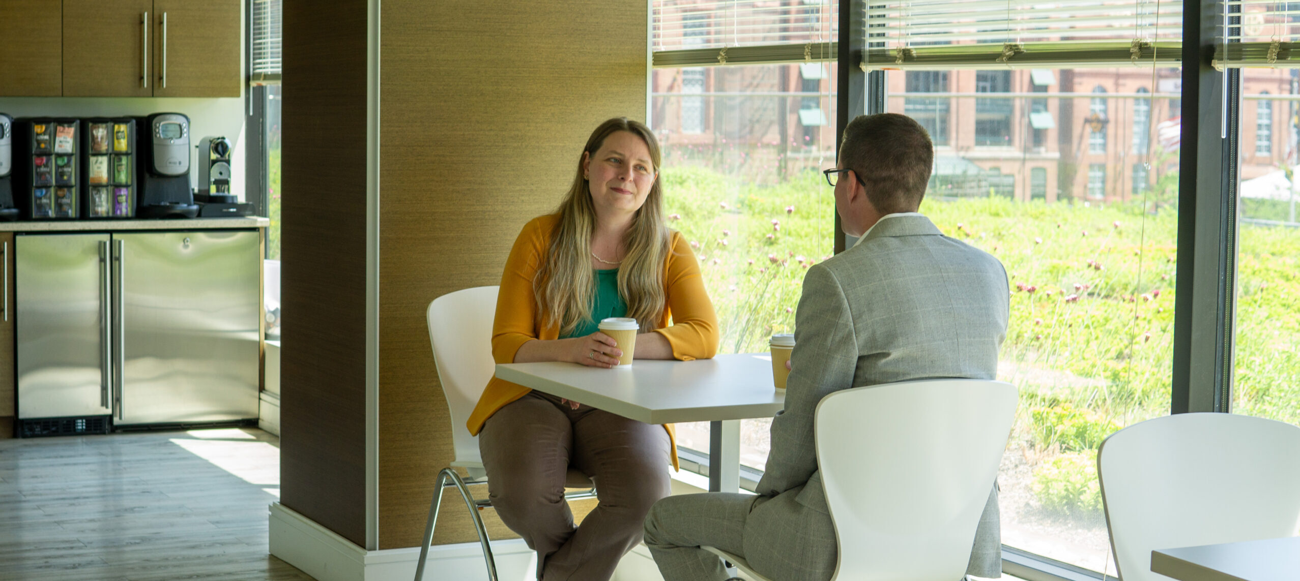 Woman in yellow sweater sits holding coffee cup across from man in a light grey suit.