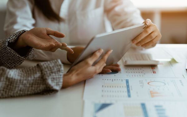 Person gestures with their hand towards a tablet being held above a table covered in documents with data.