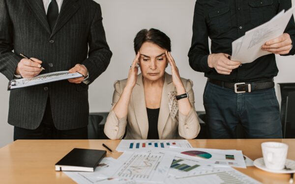 A frustrated woman in a blazer sitting at a table covered with papers with her head in her hands