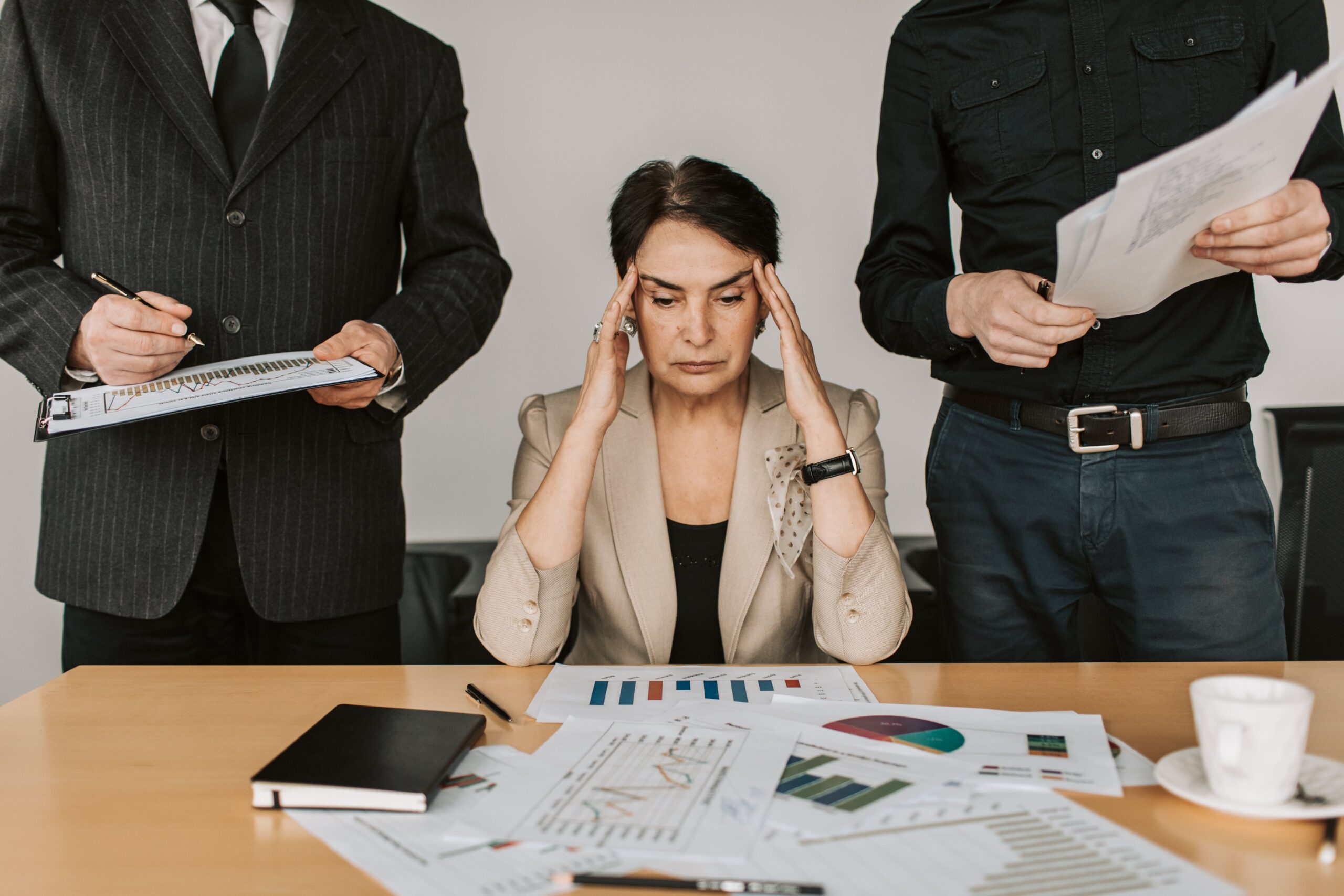 A frustrated woman in a blazer sitting at a table covered with papers with her head in her hands