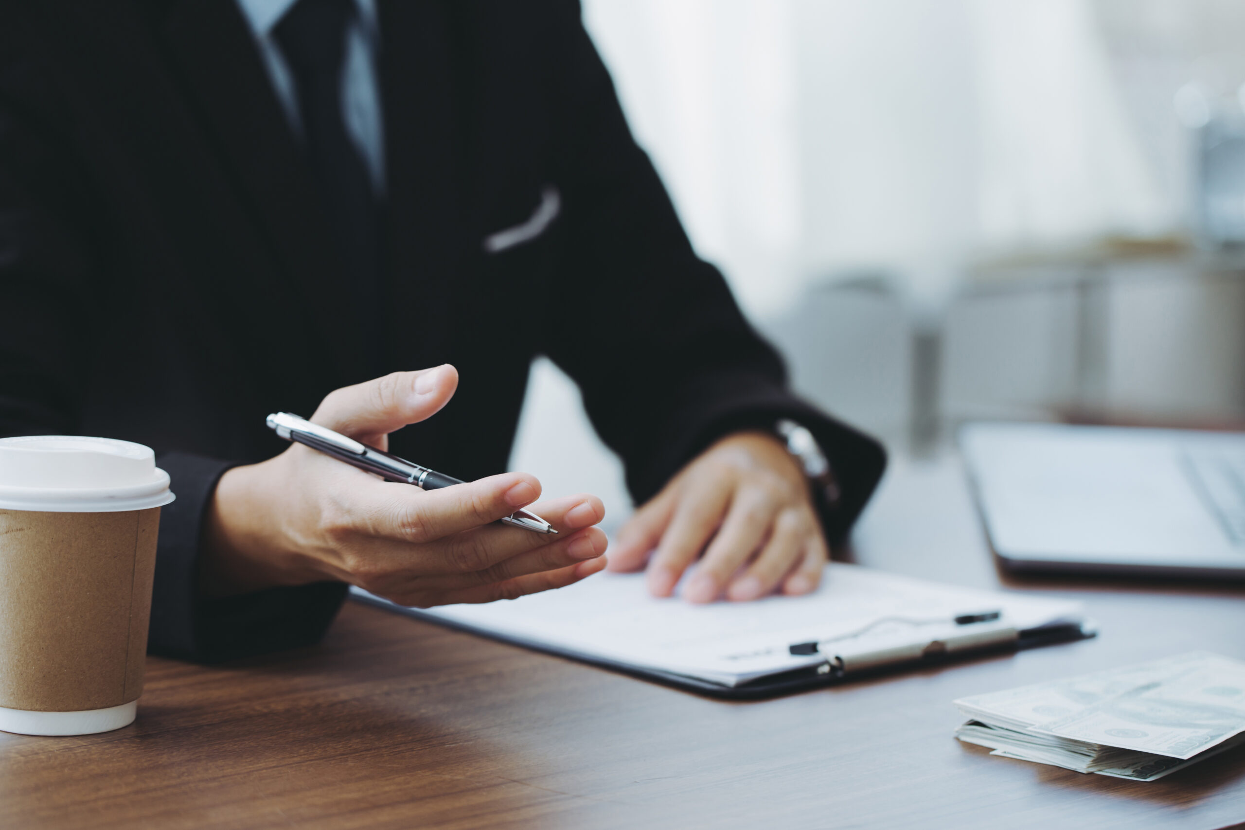 Person in suit gestures with pen in hand and clipboard on table in front of them