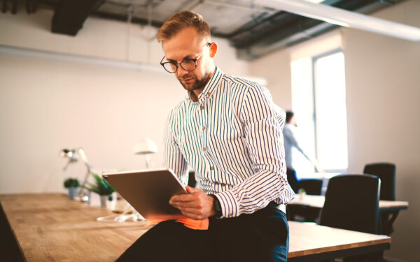 Blonde man in business attire holding tablet while sitting on desk