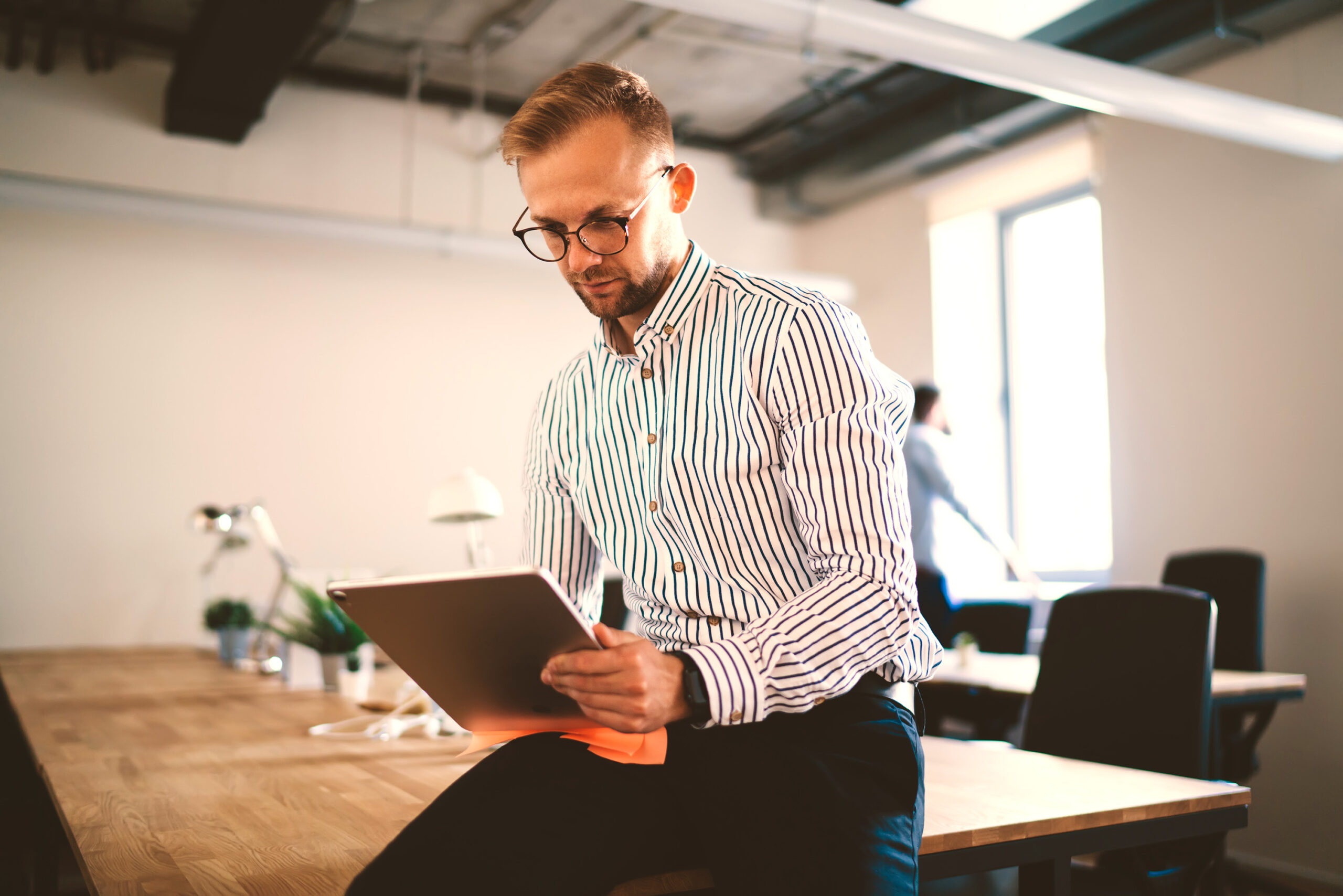 Blonde man in business attire holding tablet while sitting on desk