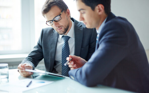 Two men in gray suits lean on a desk reviewing paperwork