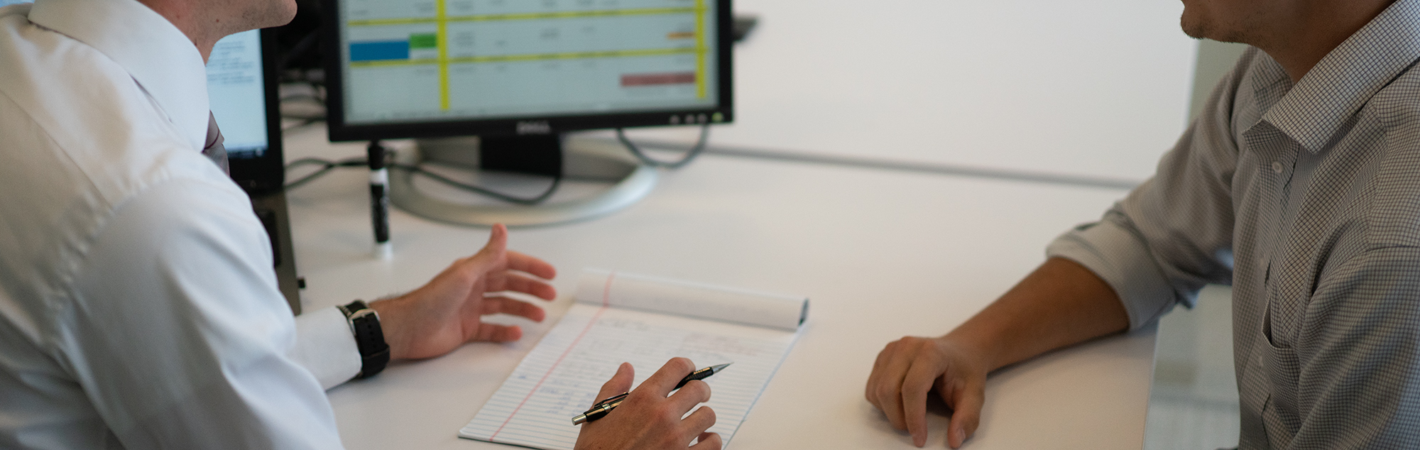 Two men in button up shirts sit at desk discussing a spreadsheet on a computer screen.
