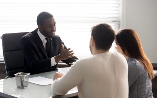 Middle aged man in black suit gives advice to couple sitting across from him at a desk in a corporate office