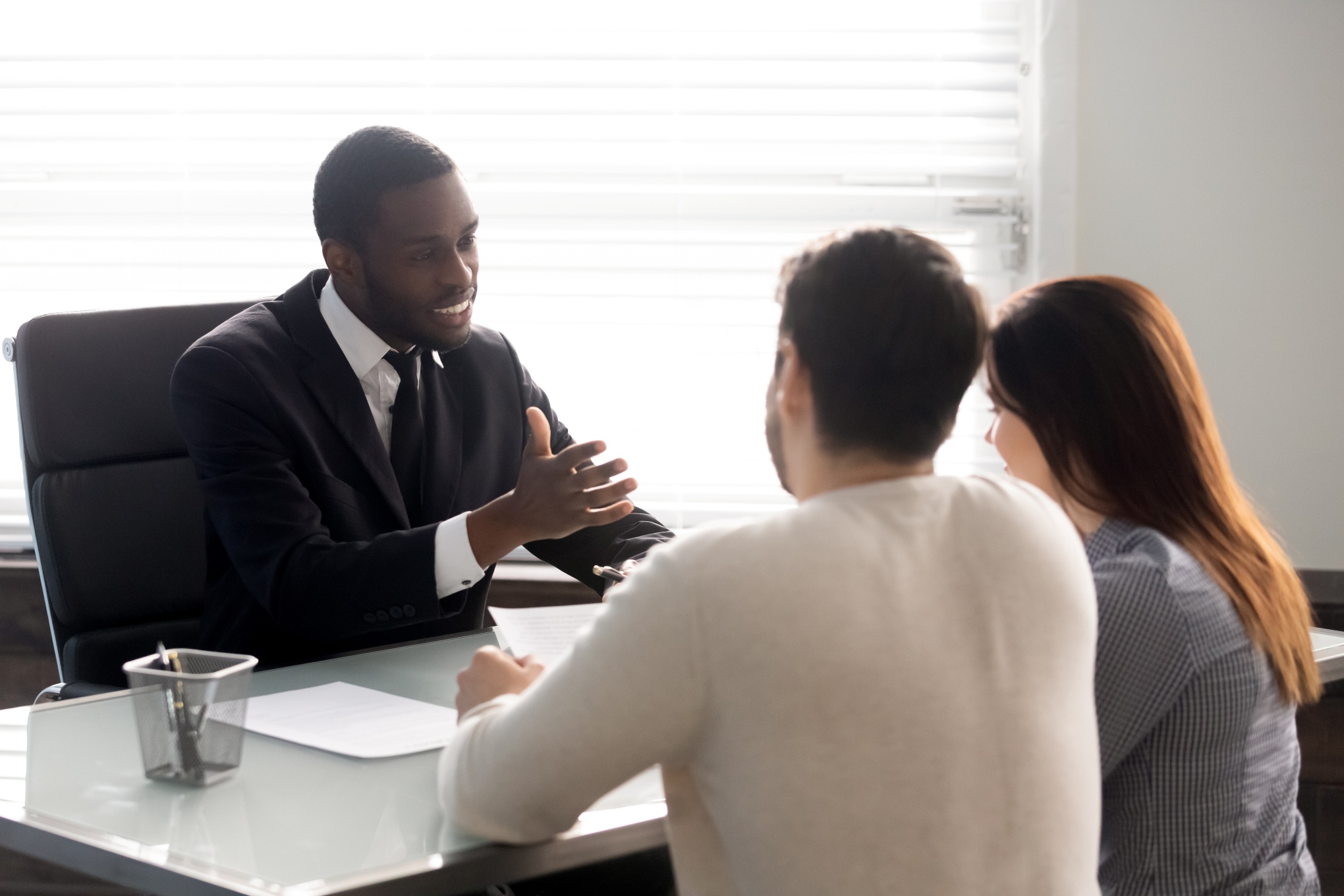 Middle aged man in black suit gives advice to couple sitting across from him at a desk in a corporate office