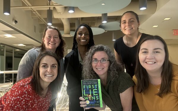 Six women pose together in a photo, one woman is holding a book titled 