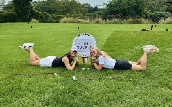 Carly Johnson and Renee Collins pose next to a sponsorship sign that says Ellin & Tucker on a golf course