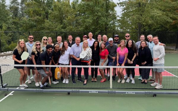 Members of Kelly Benefits Group and Ellin & Tucker stand together posing for a photo on a pickleball court on an overcast day