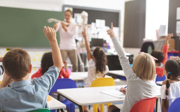 School children seated at desks raising hands in a brightly lit classroom