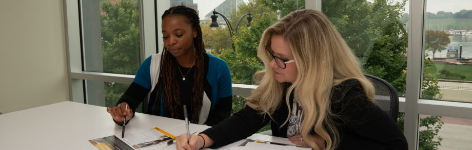 Jayla Prince and Amanda Johnston sit at a white table discussing documents with Baltimore skyline in background