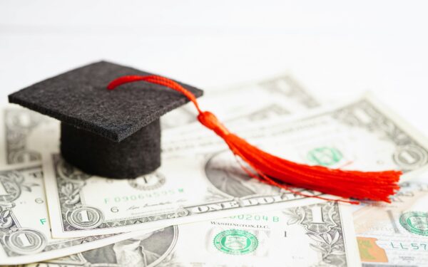 small felt black graduation cap and tassel sit on top of US dollar bills spread out on table