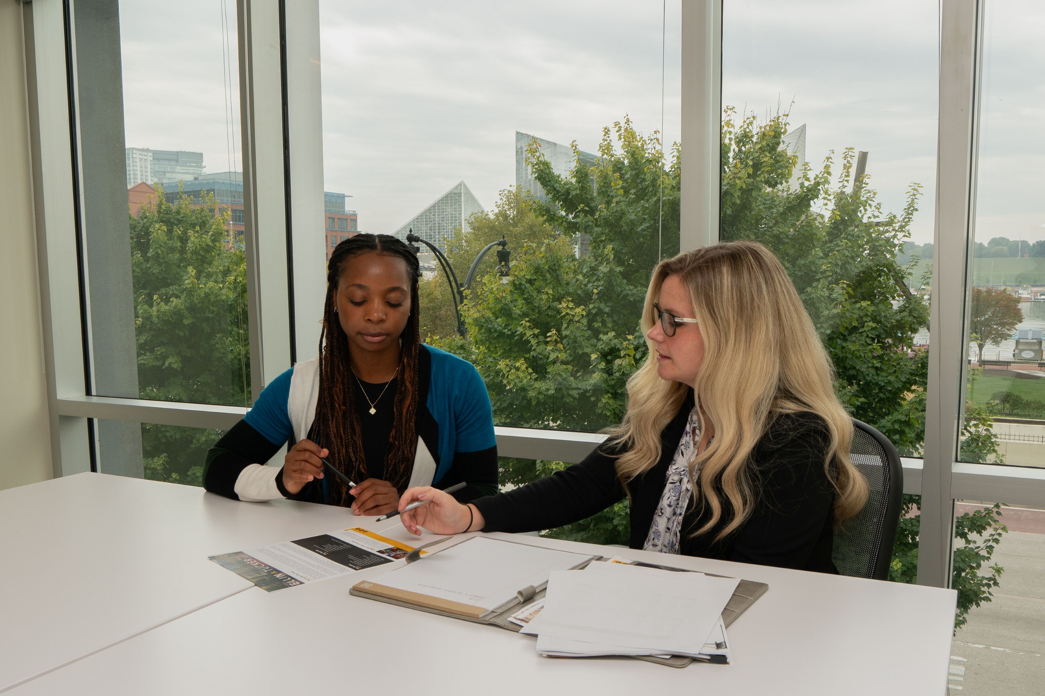 Jayla Prince and Amanda Johnston sit at white table in an office reviewing documents with the Baltimore Inner Harbor in the background