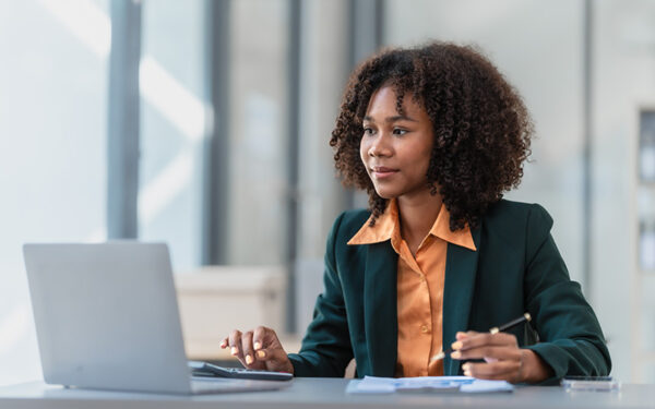 Woman in orange button up shirt and blazer sitting at desk looking at a laptop