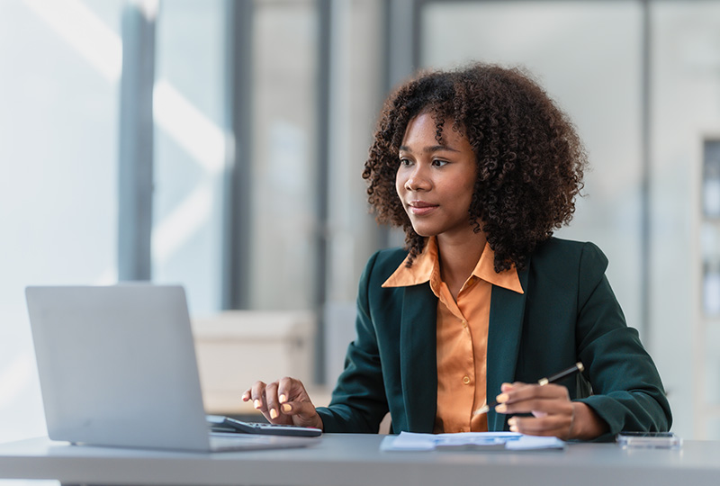 Woman in orange button up shirt and blazer sitting at desk looking at a laptop