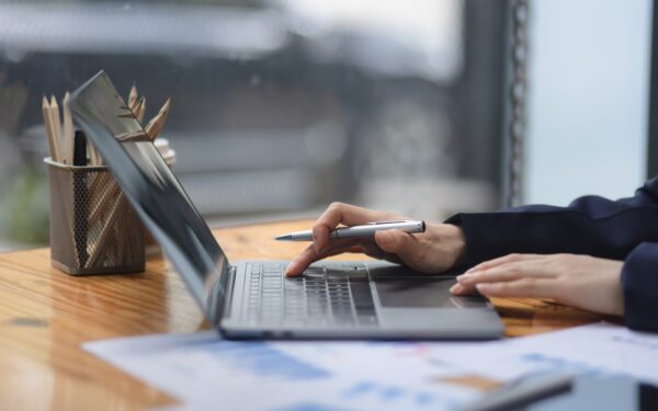 hand clicking track pad on silver laptop sitting on wooden desk next to spreadsheets