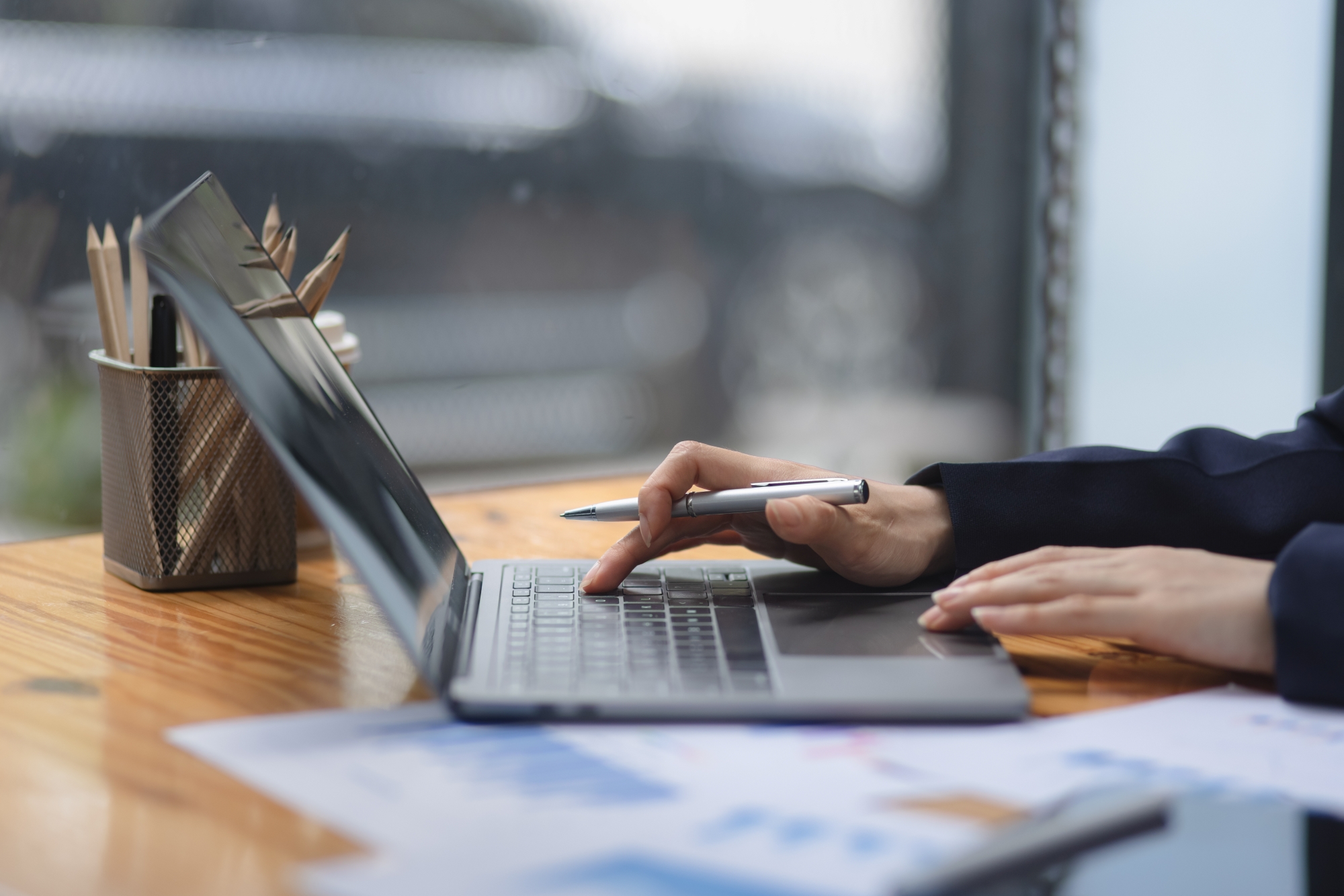 hand clicking track pad on silver laptop sitting on wooden desk next to spreadsheets