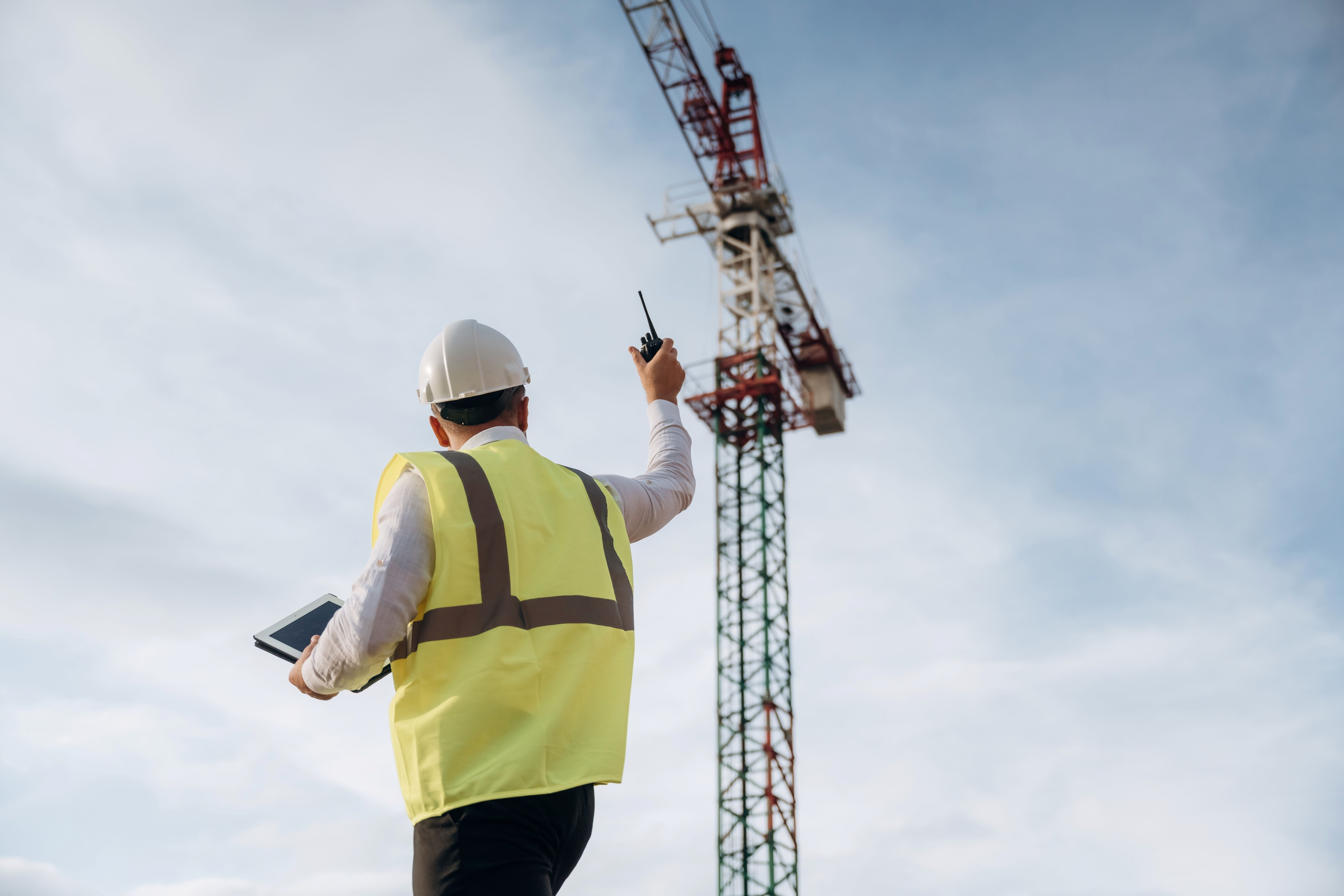 Construction worker in yellow vest and hardhat raises hand with walkie talkie up towards a crane